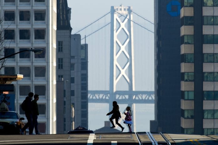 Pedestrians cross the street in front of the Oakland-San Francisco Bay Bridge in California | David Paul Morris/Bloomberg