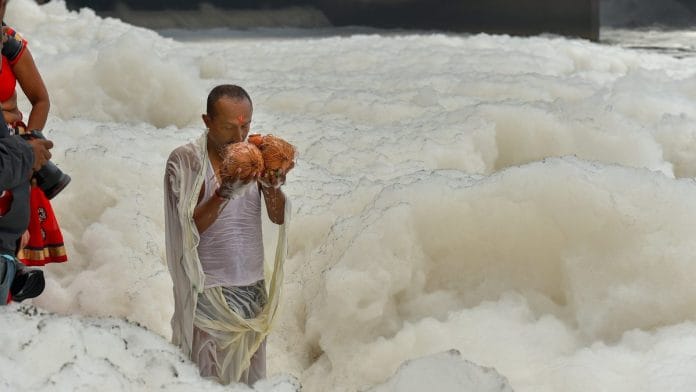 A devotee offers prayers to the rising Sun during Chhath Puja as toxic froth floats on the surface of polluted Yamuna river, at Kalindi Kunj, in New Delhi