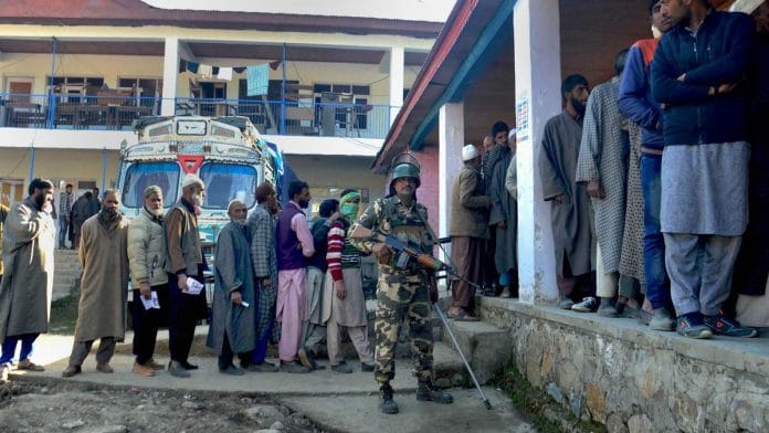 A security personnel stands guard as voters stand in a queue to cast their votes at a polling station in Anantnag district of Jammu and Kashmir