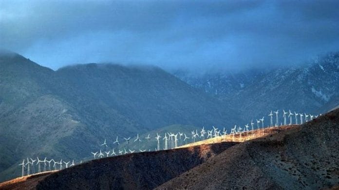 A band of sunlight shines through storm clouds to illuminate wind generators at a wind farm | Photo by David McNew | Bloomberg
