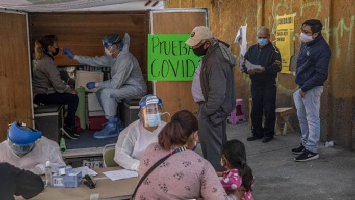 A medical worker wearing protective gear tests a resident while people stand in line at a temporary Covid-19 testing kiosk in the Pedregal de Santo Domingo, Mexico | Photographer: Alejandro Cegarra | Bloomberg
