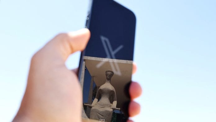 The logo of social media platform X is seen on a mobile phone next to a reflection of Alfredo Ceschiatti's sculpture The Justice, outside Brazil's Supreme Court in this illustration taken August 30, 2024, in Brasilia, Brazil | Reuters