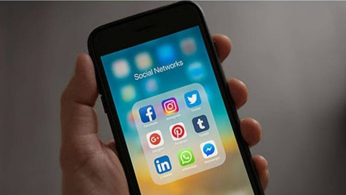 A hand holding a phone that displays social media apps such as Facebook, Instagram, X, and WhatsApp