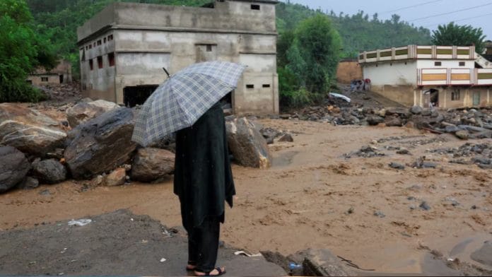 A resident stands with an umbrela as rain water flowing from mountains crosses a damaged area, following a storm that caused heavy rains and flooding in Bayshonai Kalay, in Buner district, in Khyber Pakhtunkhwa province, Pakistan, August 18, 2025. | REUTERS