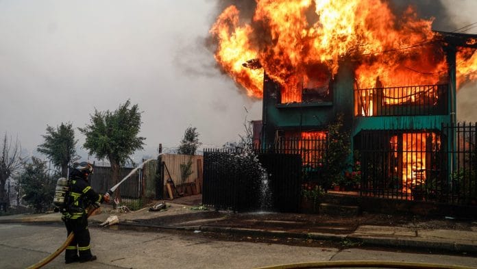 A firefighter tries to extinguish a fire in front of a burning building as fire and smoke rise from a forest fire in the Biobio region where, according to local media, multiple wildfires prompted emergency evacuations, in Concepcion, Chile, January 18, 2026 | Reuters