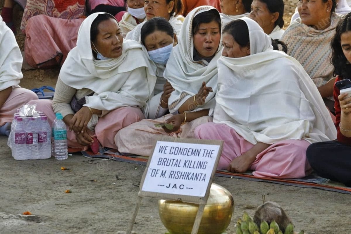 Dozens of women sit beneath a tree, wearing white shawls and pink phaneks (wrap-around skirts). Posters read: 'We condemn the brutal killing of M. Rishikanta.' | Praveen Jain, ThePrint