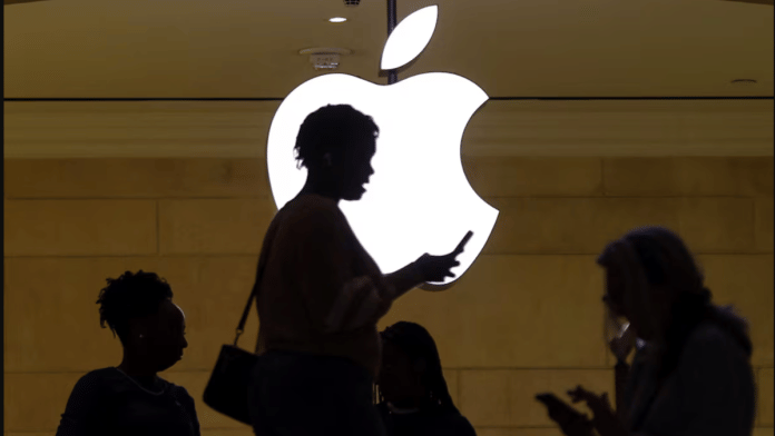 A woman uses an iPhone mobile device as she passes a lighted Apple logo at the Apple store at Grand Central Terminal in New York City, U.S., April 14, 2023. | REUTERS/Mike Segar