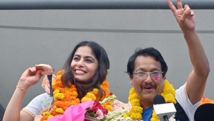 New Delhi, Aug 07 (ANI): Double Olympic medalist Manu Bhaker shows her medal as she arrives in New Delhi on Wednesday, after her historic performance in the Paris Olympics 2024. She won bronze medals in Women’s 10m Air Pistol and 10m Air Pistol Mixed team event. Her coach Jaspal Rana also seen. (ANI Photo/Sanjay Sharma)