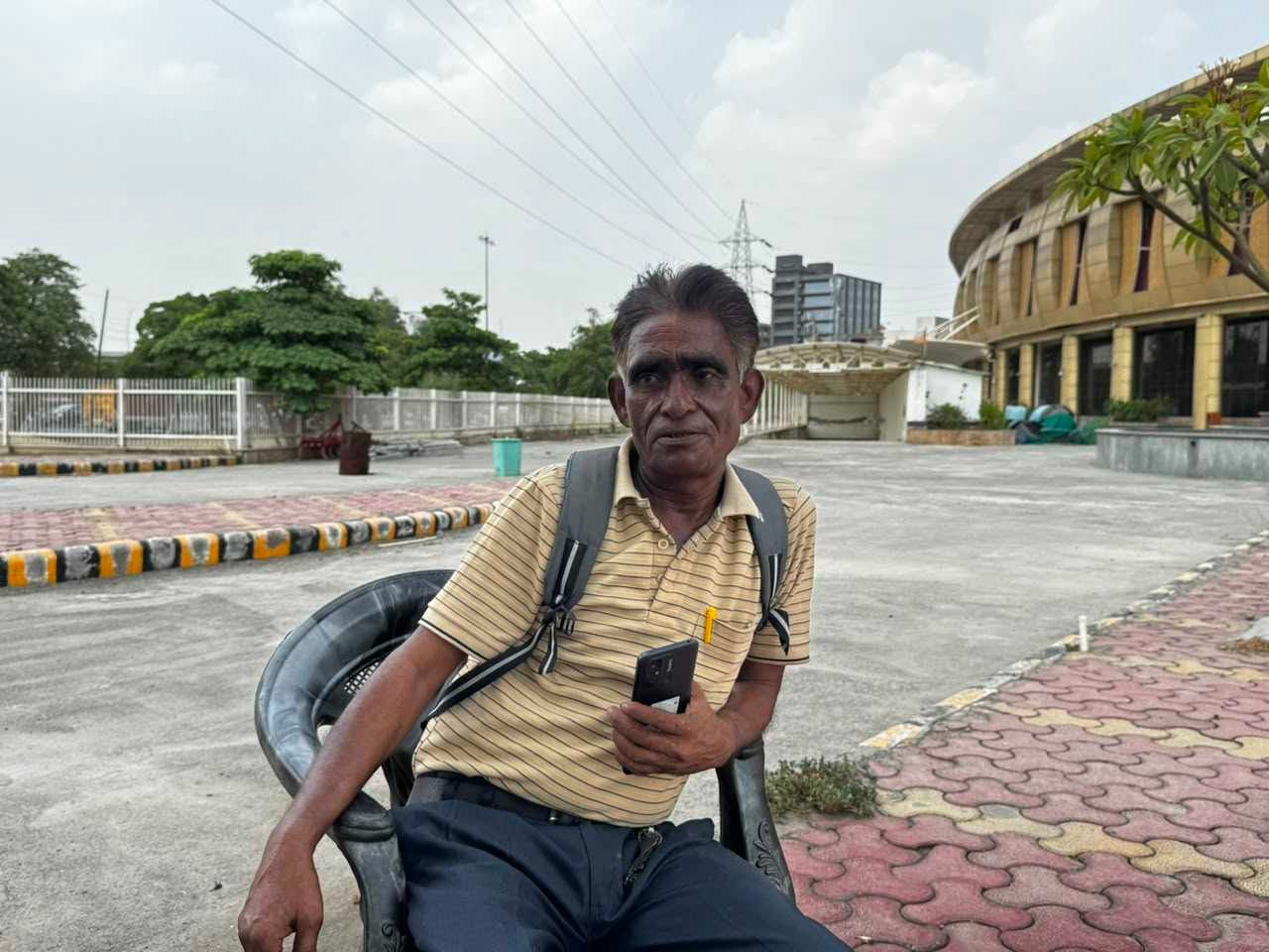 An elderly man sits in a plastic chair with the bus terminal at his back.