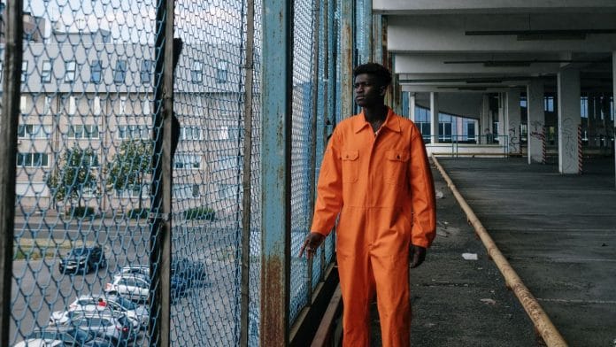 An adult male in an orange jumpsuit stands by a wire mesh fence in an urban setting indoors.